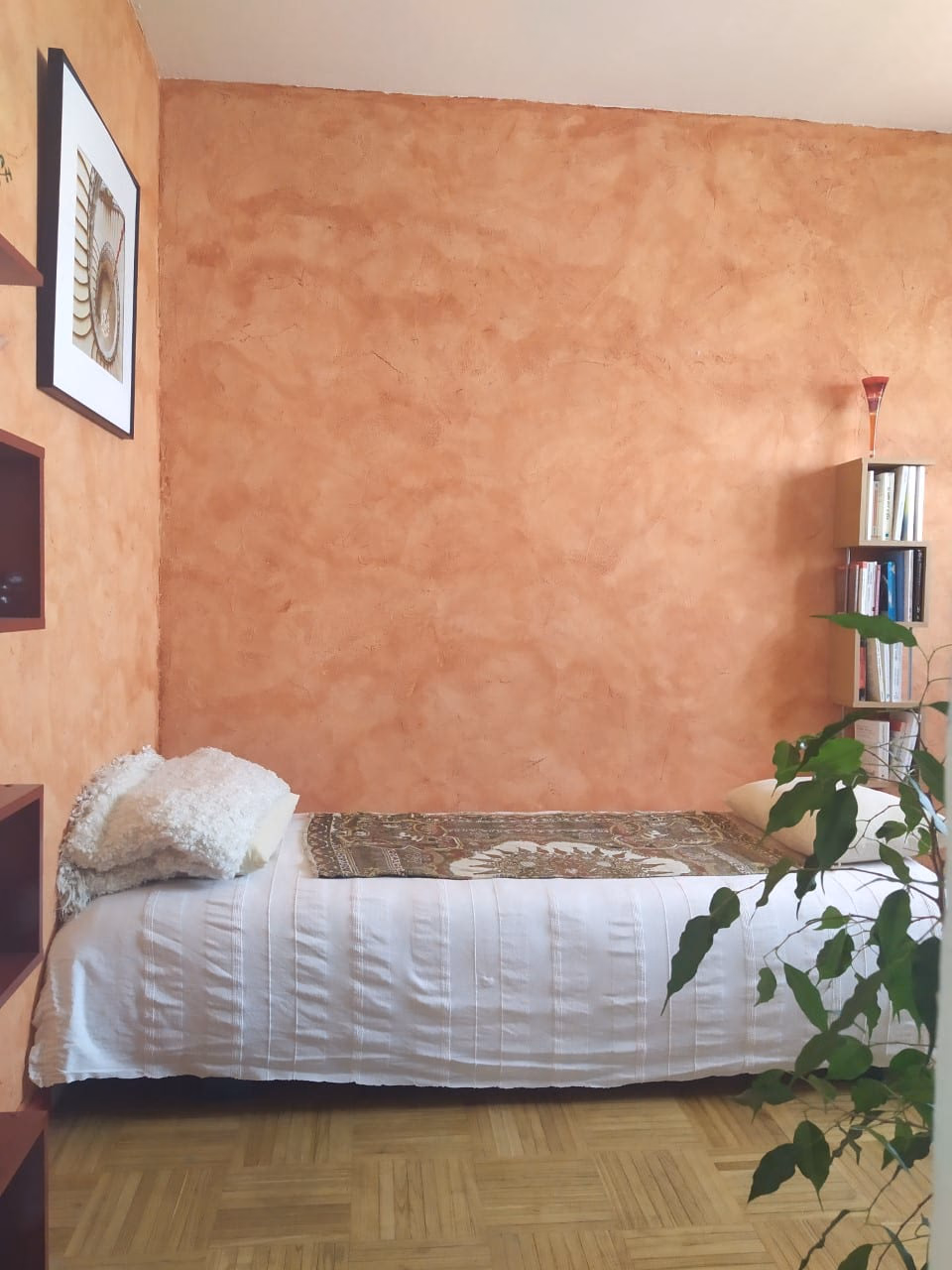 Un divan recouvert d'un tissu blanc et d'un châle à motifs, contre un mur ocre, à côté d'une petite bibliothèque et d'une plante verte.